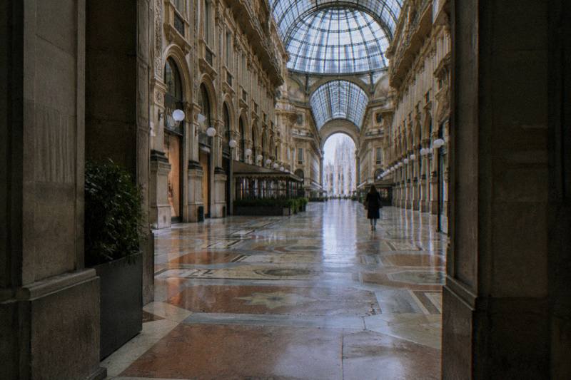 Morning inside the Galleria Vittorio Emanuele II in Milan, Italy, an on-foot walking tour seen from under an arched doorway along the mosaic pavement, iron-and-glass roof filtering pale light, a scuffed marble floor and one distant figure from behind.