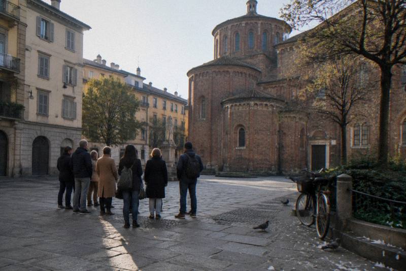Early morning at Largo Ildefonso Schuster in the Ticinese district of Milan, Italy, a small tour group gathered on the small square's worn paving, low sun raking across weathered brick and stone facades, a parked bicycle and scattered pigeon feathers near the square's edge.