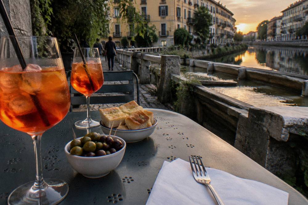An aperitivo spread of drinks and snack bowls on an outdoor table beside a canal