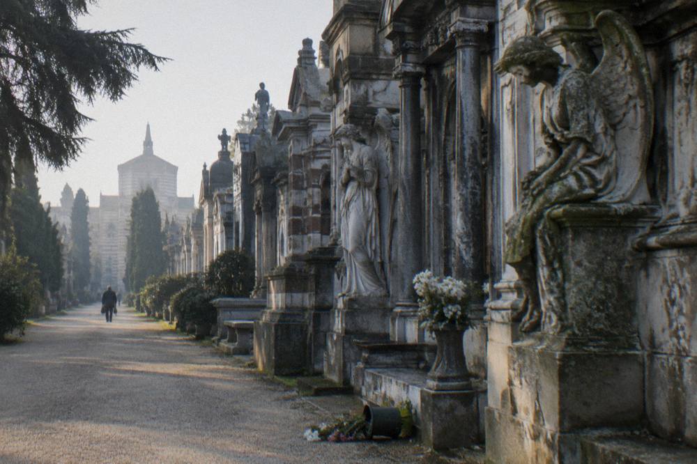 Row of ornate stone family tombs with carved angels and columns along a cemetery avenue