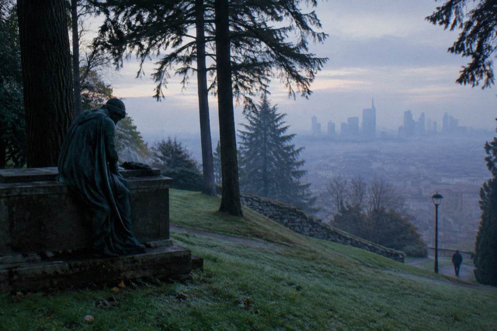 Sculpted bronze mourner figure beside a stone tomb under tall trees