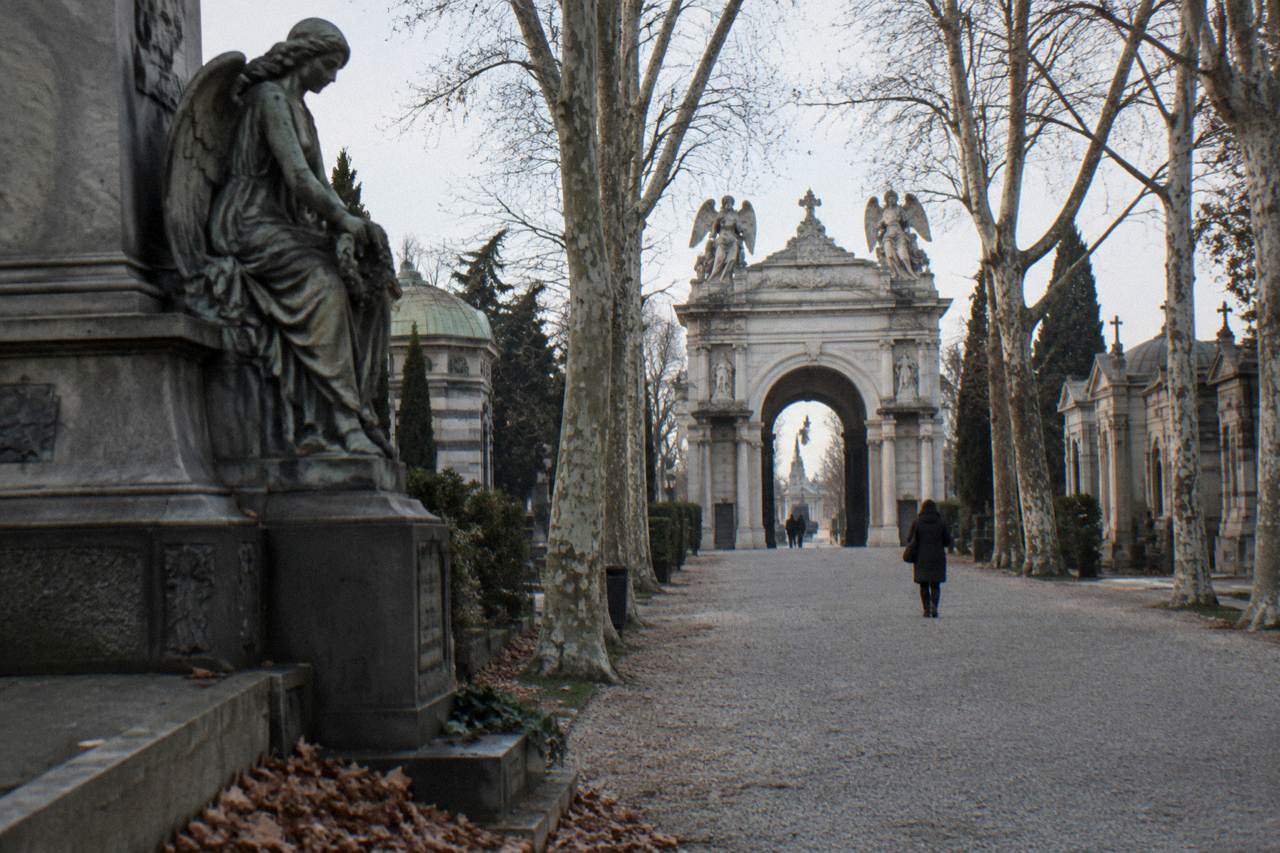 Ornate cemetery facade with carved stone arches and sculpted angels along a tree-lined path
