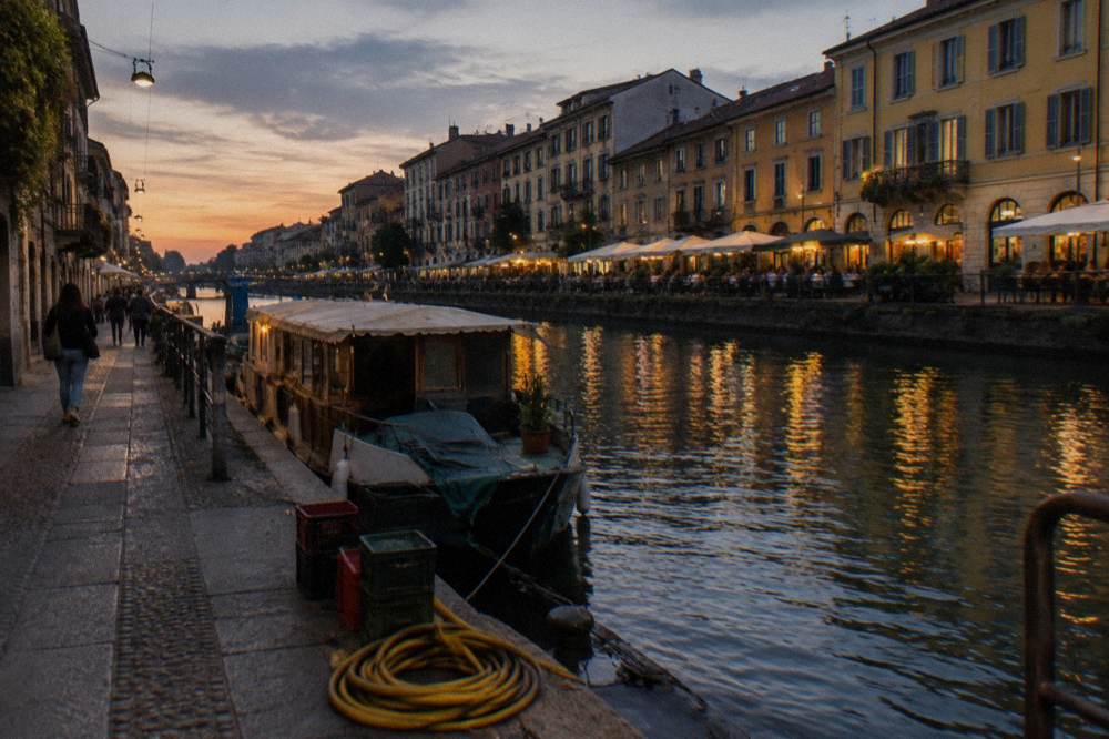 Canal-side promenade along the Naviglio Grande in the early evening with restaurant lights reflecting in the water