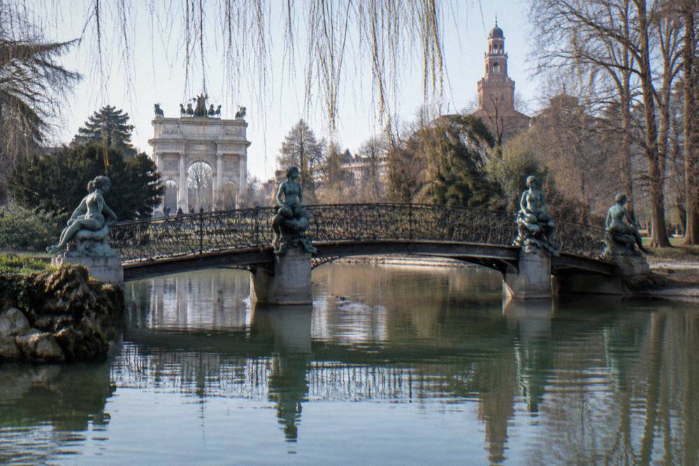 Ornate iron footbridge over a small lake in a city park with sirens statues at each corner