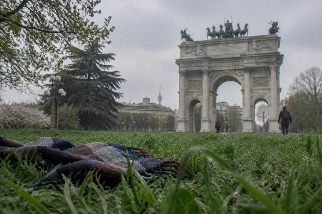 Wide green lawn of a city park with trees and a tall stone arch in the background