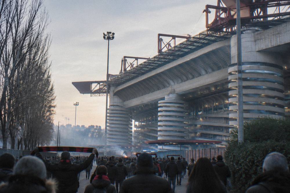 The red steel roof girders and cylindrical towers of the San Siro stadium seen from the surrounding street