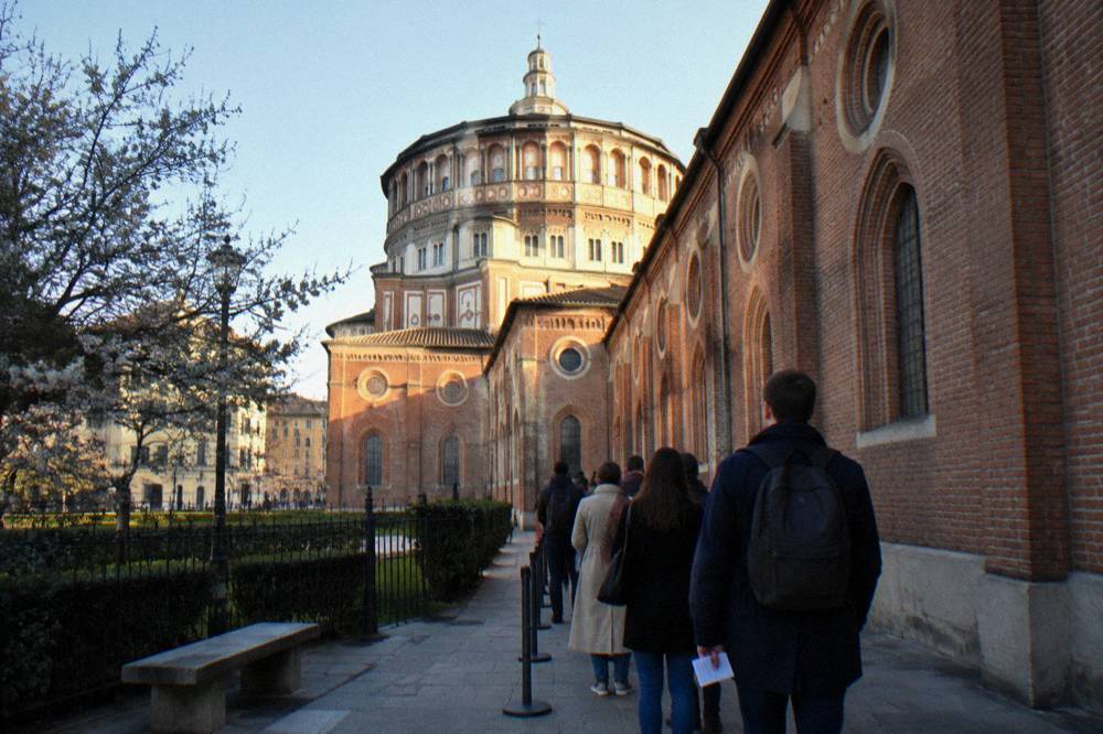 A timed-entry queue outside the brick facade of Santa Maria delle Grazie in Milan on a clear afternoon