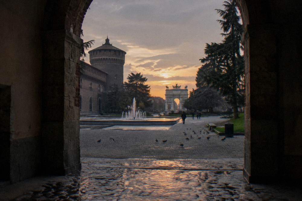 The courtyard and fountain of the Sforza castle in Milan with the Arco della Pace visible through the archway at dusk