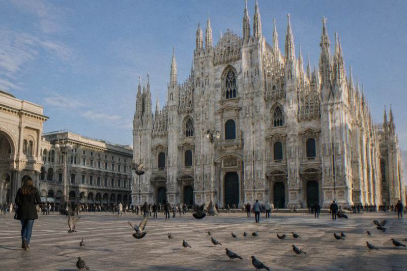 Visitors cross the vast open expanse of Piazza del Duomo in Milan, the cathedral's white marble facade filling the frame behind them under a clear sky.
