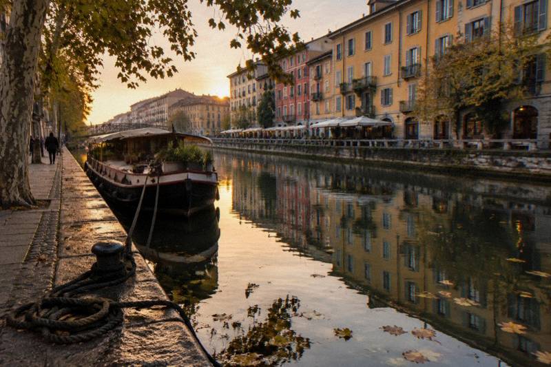 A quiet Navigli side street at dusk, bicycles leaning against faded shutters and canal water reflecting lit windows.