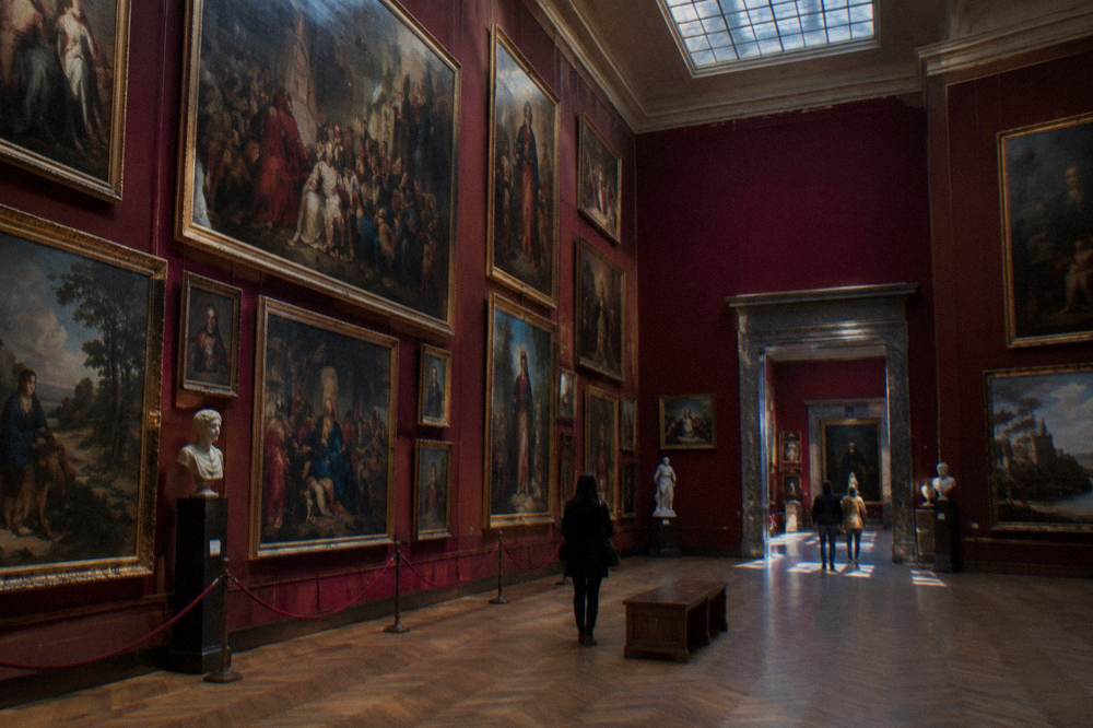 Long gallery hall with paintings in ornate frames hung salon-style on a wall lit by skylights
