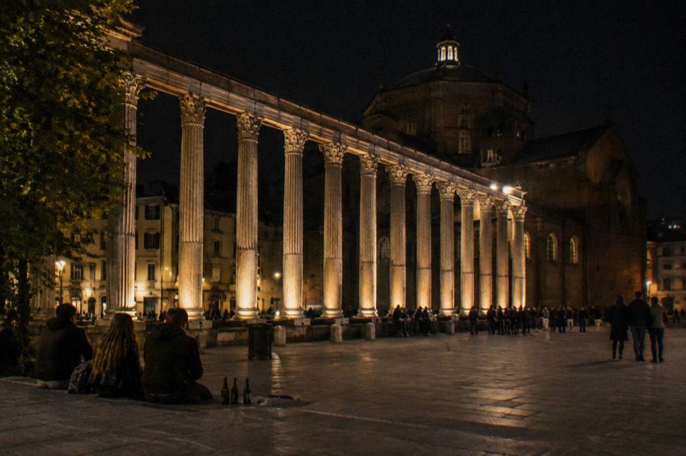 The Roman columns of San Lorenzo in Milan lit by warm lamps with people gathered in the square