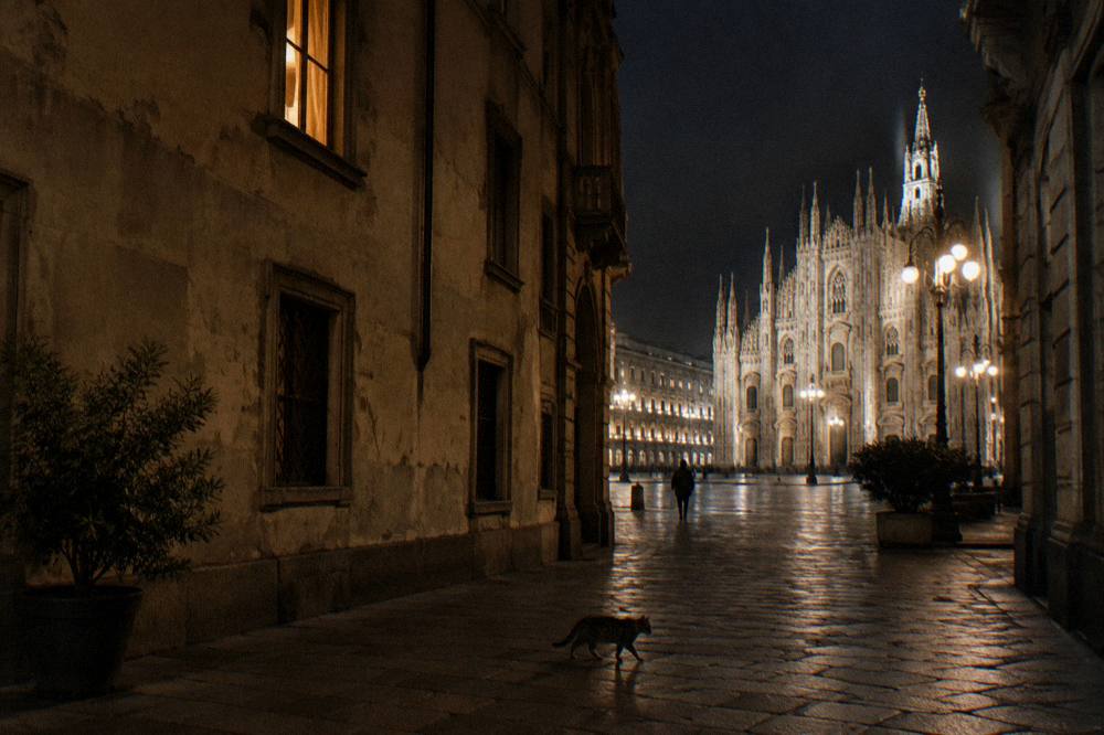 A lamplit Milan street at night with the Duomo spires visible above the rooftops
