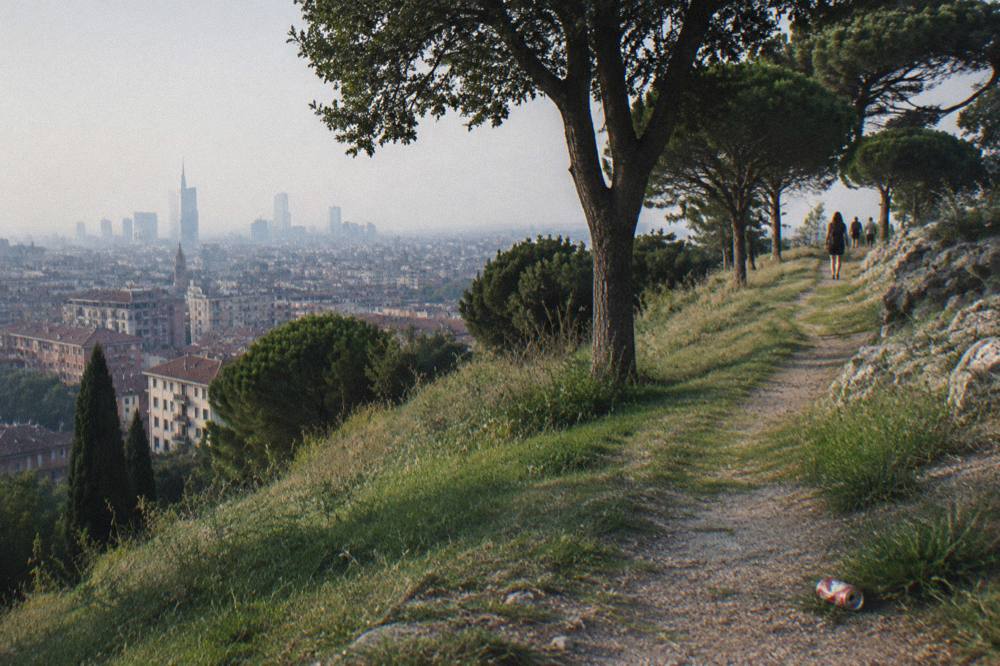 A green hillside park path above city rooftops, with trees, grass and distant apartment blocks under a wide sky